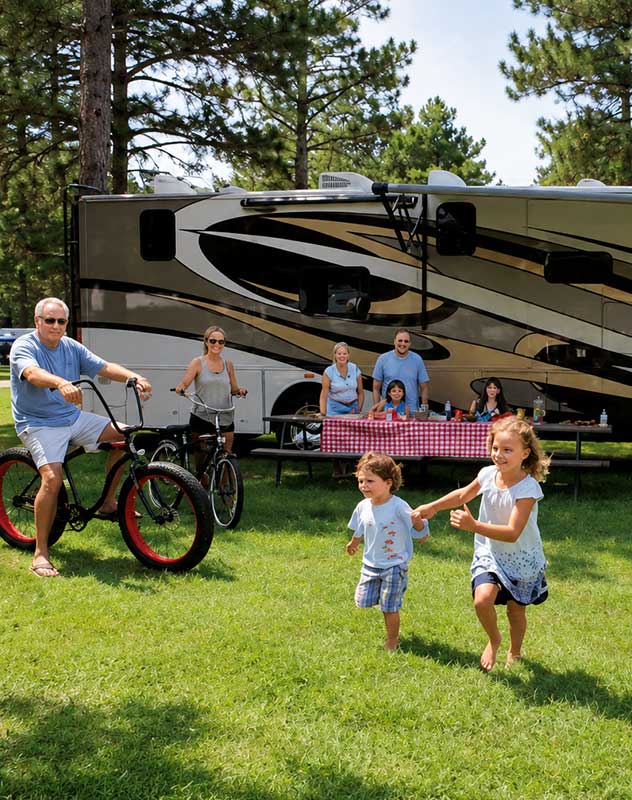 A happy family sitting in lawn chairs outside their luxury RV camper at a private resort, watching their kids run and play on a spacious grassy campsite.