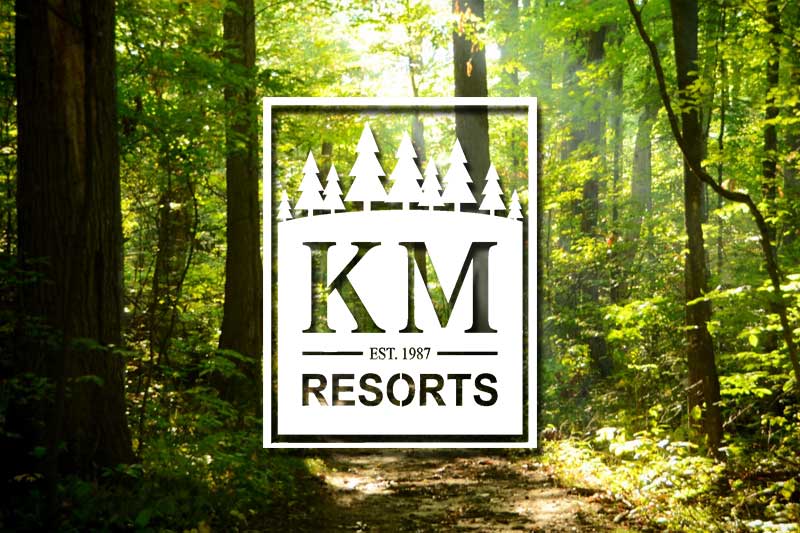 A premium travel trailer RV camper parked on a spacious campsite next to a calm lake at a K/M Resorts of America private campground preserve in the Pacific Northwest.