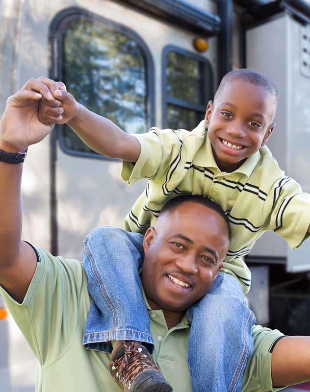 Parents relaxing by their fifth-wheel RV trailer at a shaded private campground, watching their young children play tag on a manicured grassy lawn.